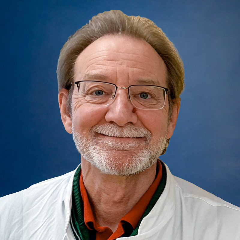 Portrait of a smiling man with glasses and a white beard wearing a white coat in front of a blue background.