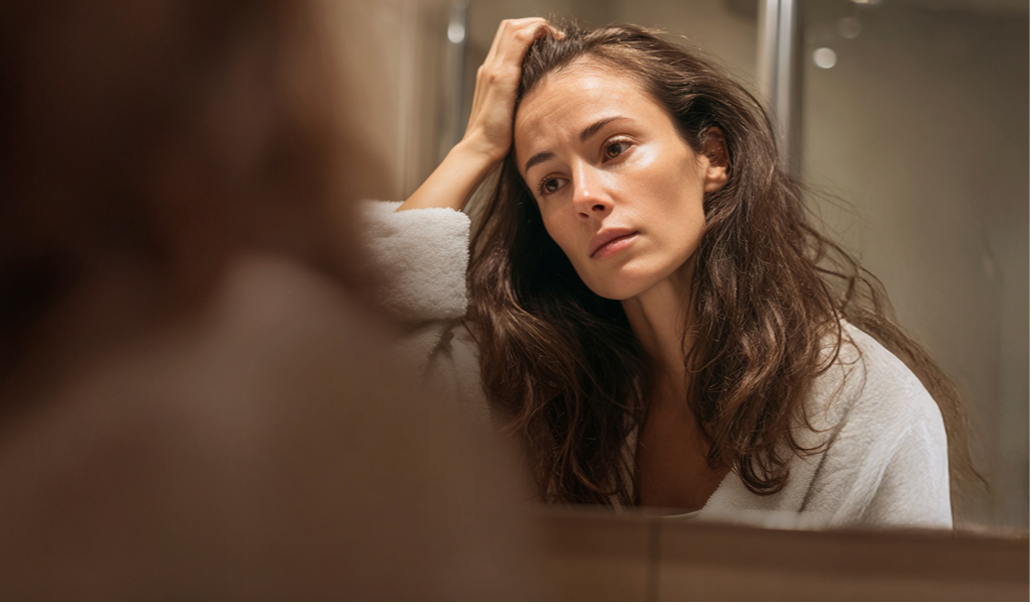 Frau im Badezimmer blickt in den Spiegel und fasst sich ins Haar, mit besorgtem Gesichtsausdruck