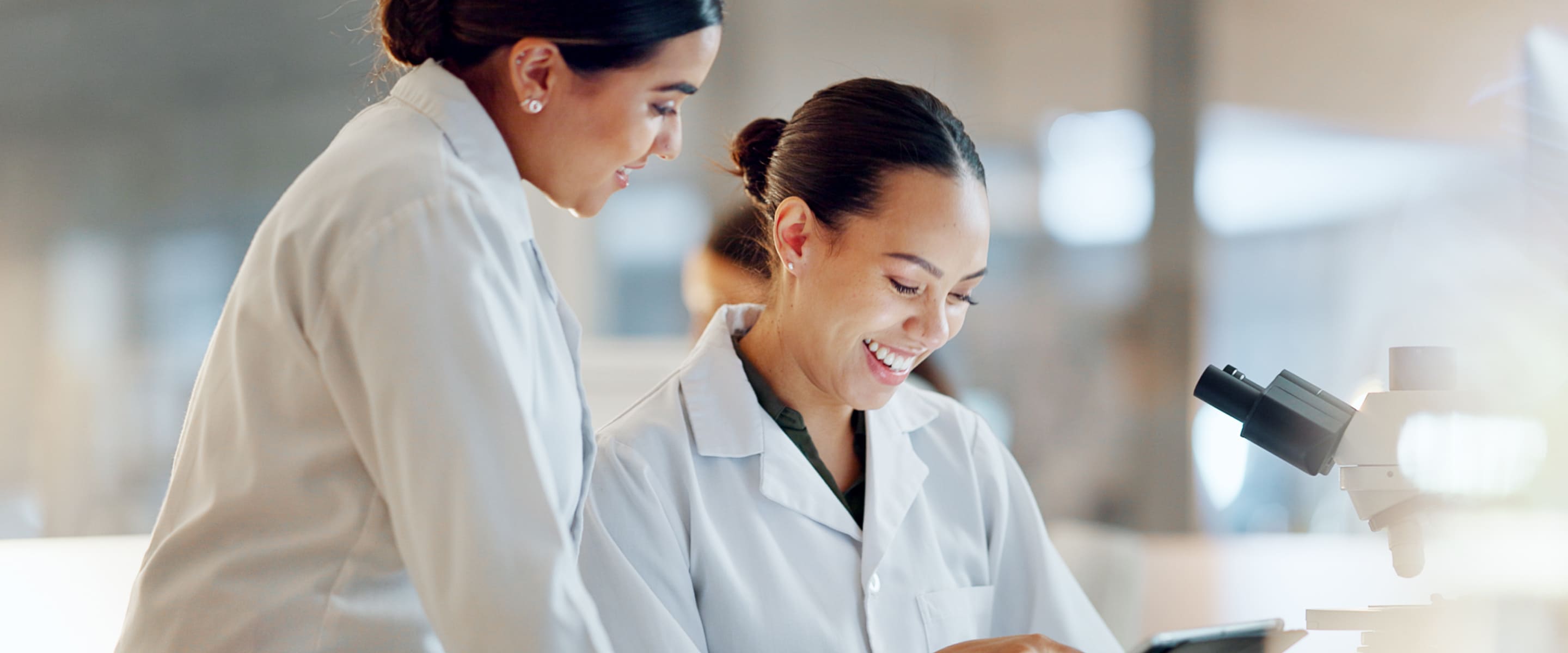 Two researchers in the laboratory smile at data on a tablet; microscope in the foreground