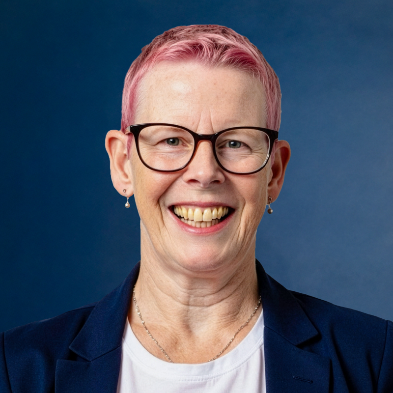 Portrait of a laughing woman with short pink hair and glasses wearing a dark blue blazer in front of a blue background.