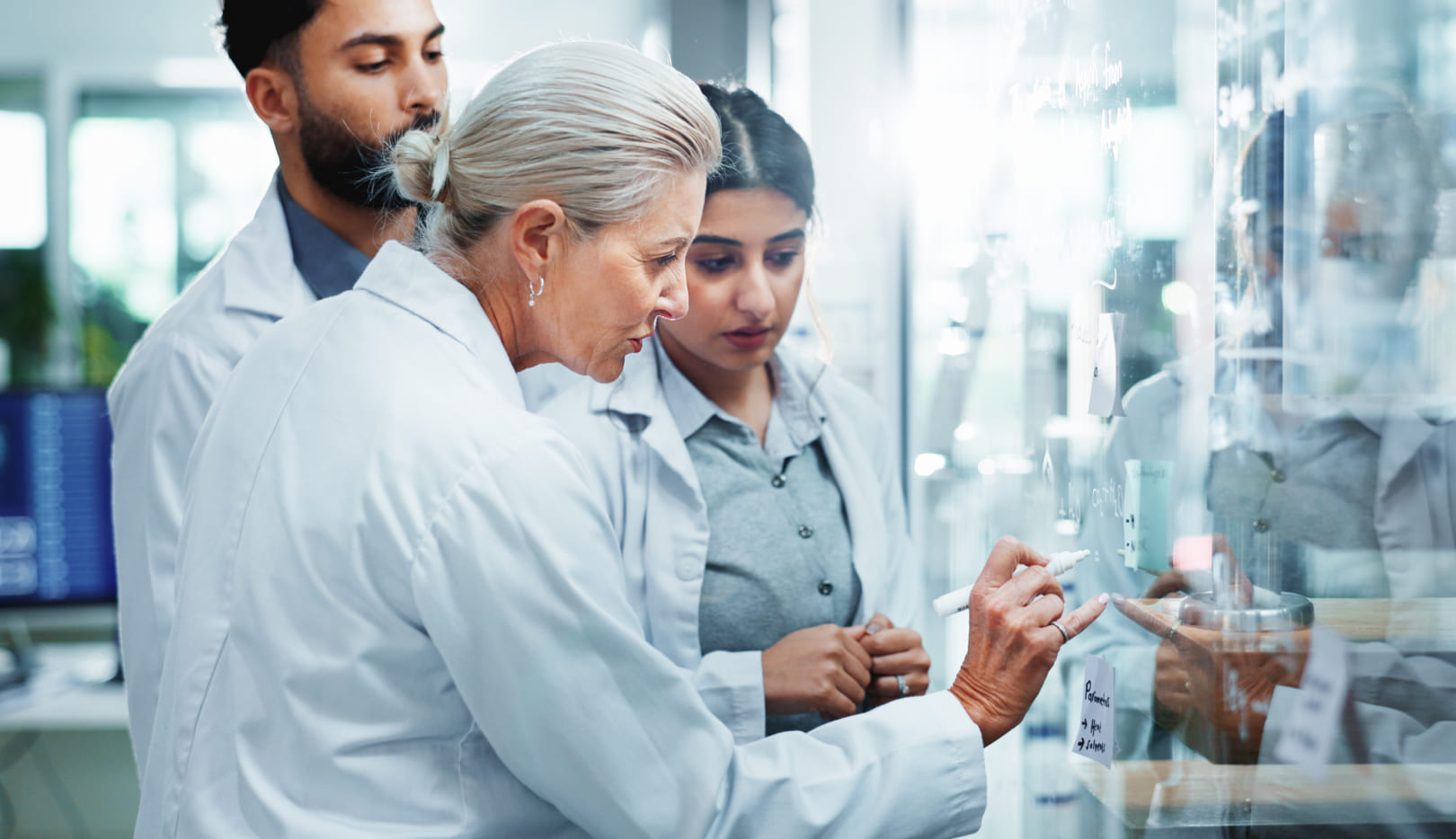 Experienced scientist explains formulas on glass wall—two younger colleagues listen