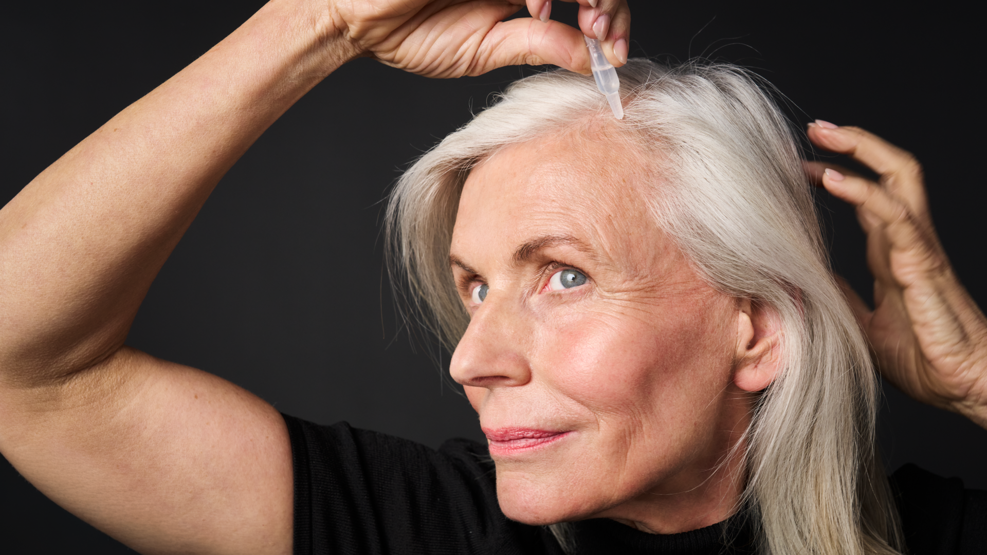 Ältere Frau mit langem, silbergrauem Haar trägt mit einer Pipette ein Haarserum auf ihren Ansatz auf