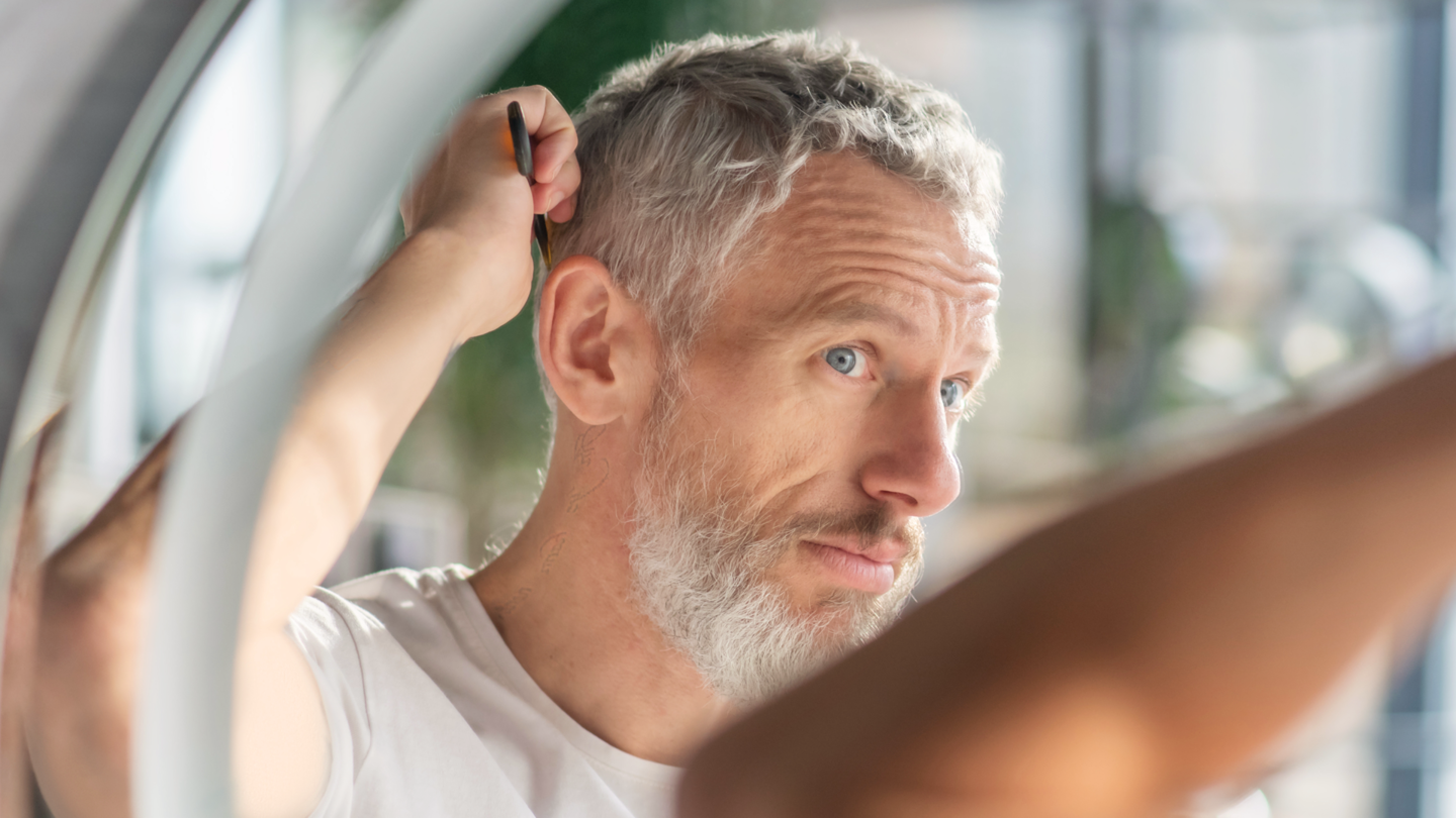 Middle-aged man with short gray hair assessing his hair fullness in front of a mirror.