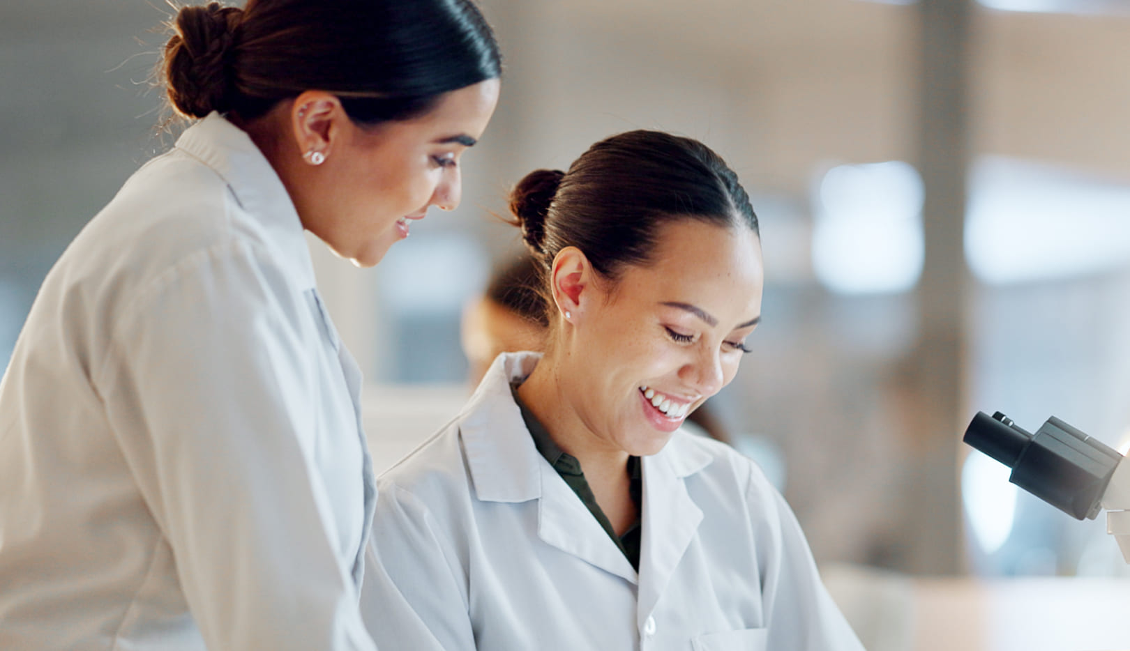 Two researchers in white lab coats smile and look together at a device next to a microscope