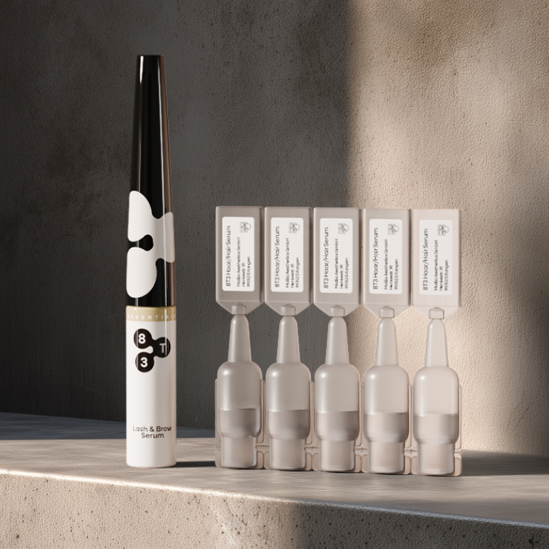 Still life: Eyelash & brow serum bottle next to five hair serum ampoules on a concrete shelf in side sunlight