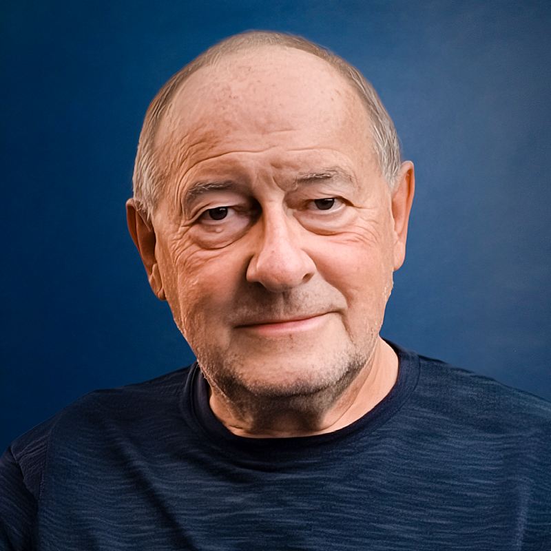 Portrait of an older man with short hair wearing a dark blue shirt in front of a blue background.