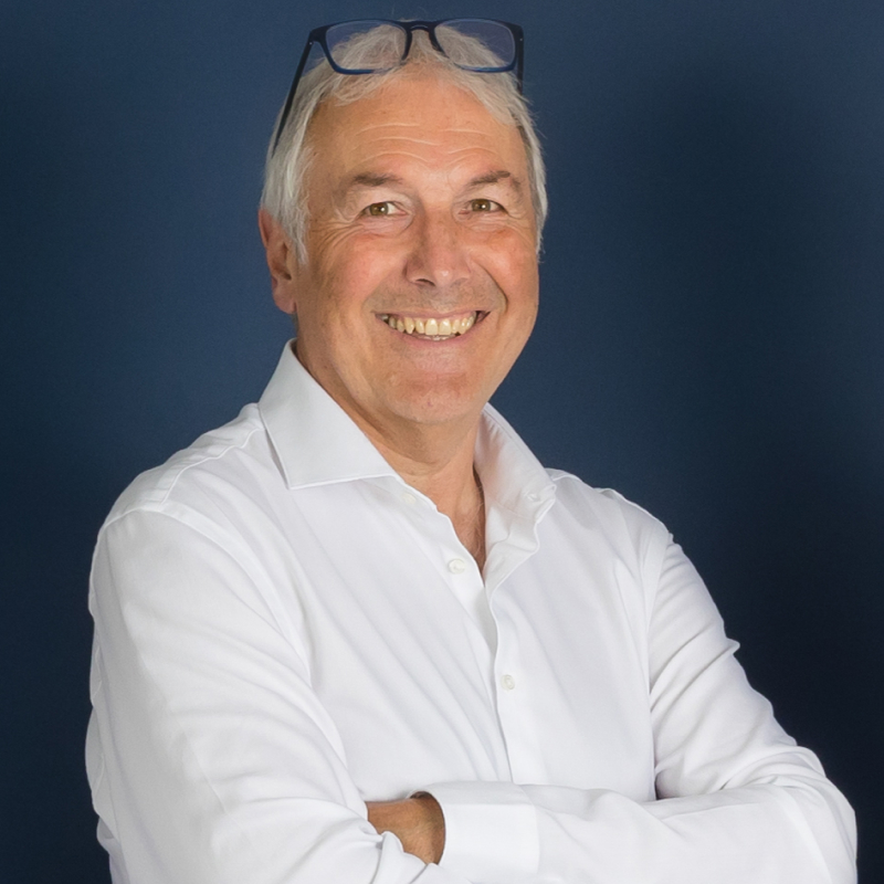 Portrait of an older man in a white shirt, smiling, blue wall, glasses on his head.