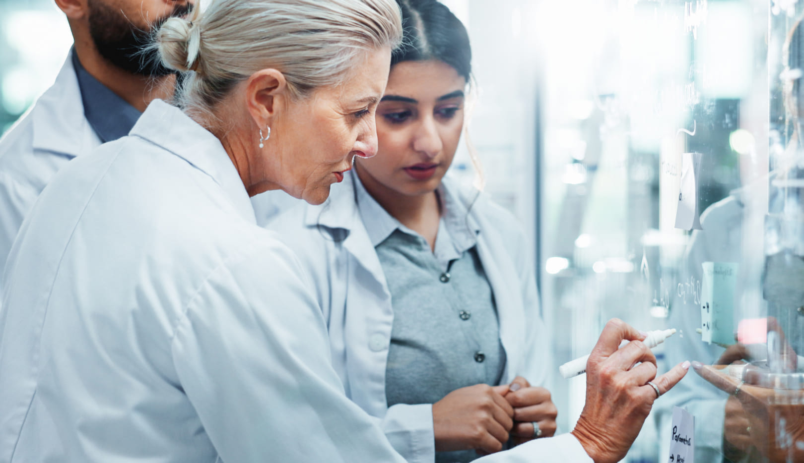 Scientist explaining formulas on a glass wall; younger colleague listening attentively